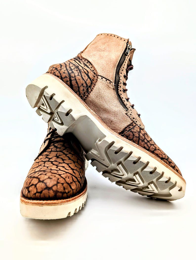 May include: Brown and tan leather boots with a unique textured pattern. The boots have a side zipper, lace-up closure, and a thick white sole. The soles have a tread pattern for grip. The boots are set against a white background.