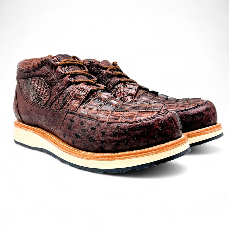 May include: A pair of brown leather boots with a textured crocodile and ostrich pattern. The boots have brown laces, a white midsole, and a light brown outsole. The boots are designed for work or casual wear.