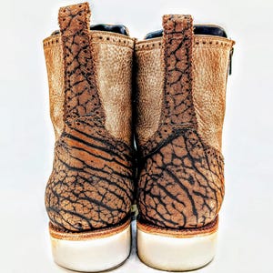 May include: Pair of brown leather ankle boots with a textured design on the back and heel. The boots have a white sole and a pull tab at the top. The leather has a distressed appearance.
