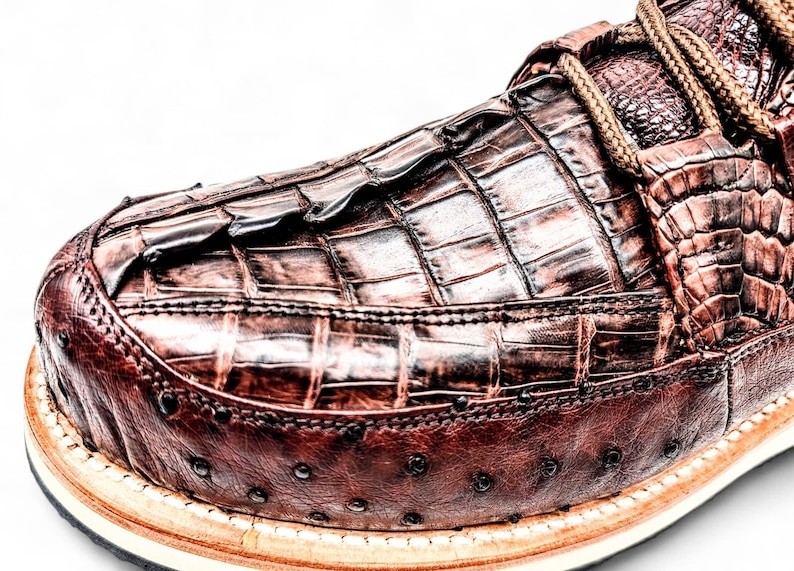 May include: Close-up of a brown leather shoe with a crocodile skin texture. The shoe features a textured vamp, decorative stitching, and a light-coloured sole. The laces are a light brown colour.