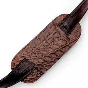 May include: Brown guitar strap with a textured, crocodile-like pattern on the shoulder pad. The strap is made of leather and has a dark brown color. The design is a close-up shot of the strap, showing the details of the material.