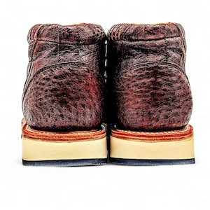 May include: A pair of brown leather boots with a textured finish. The boots feature a stacked sole with a cream base and a dark brown trim. The boots are set against a white background.