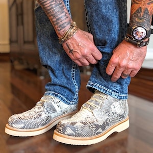 May include: A pair of brown and white snakeskin boots with white laces. The boots have a thick white sole and are being worn by someone in blue jeans.