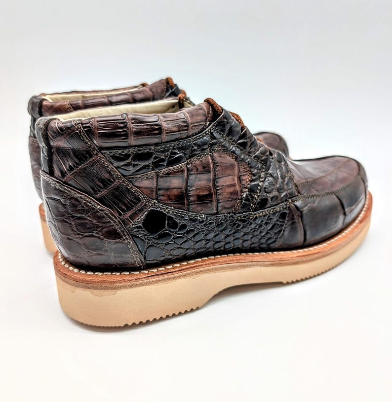 May include: Brown leather boots with a crocodile skin texture. The boots have a light brown sole and a dark brown upper with a textured pattern. The boots are designed with a classic style.