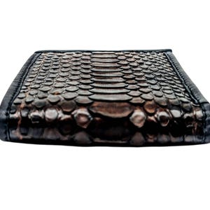 May include: A rectangular wallet with a snakeskin pattern in shades of brown and black. The wallet has a textured surface and a black border. The design is detailed and the overall look is luxurious.