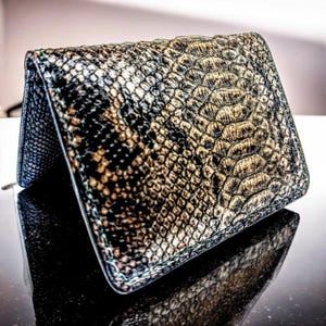 May include: A bi-fold wallet featuring a textured snakeskin pattern in shades of black, brown, and metallic gold. The exterior displays a repeating scale design with a glossy finish, showing a folded structure with visible interior edges.