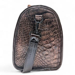 May include: A brown duffel bag with a crocodile skin texture. The bag has a rounded shape with a dark brown leather strap and a black metal clasp. The bag is designed for travel and has a luxurious appearance.