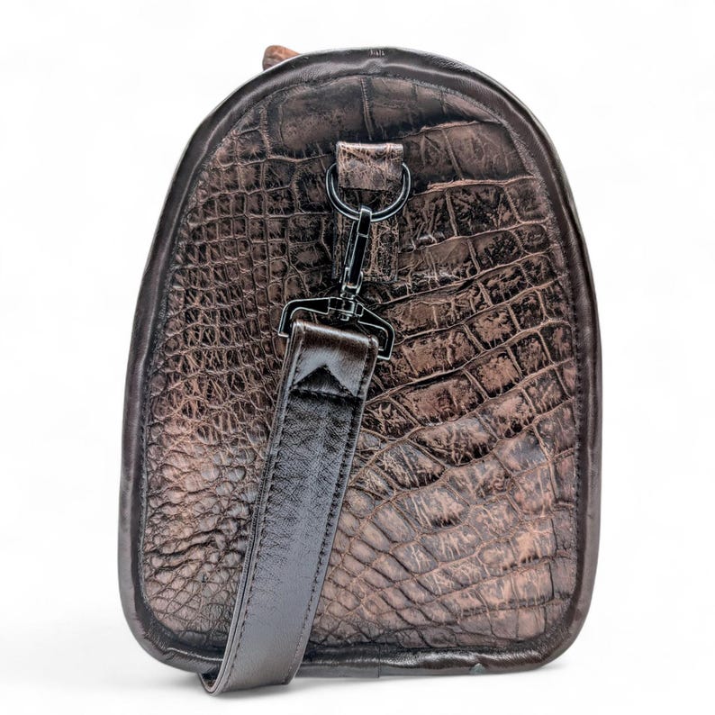 May include: A brown duffel bag with a crocodile skin texture. The bag has a rounded shape with a dark brown leather strap and a black metal clasp. The bag is designed for travel and has a luxurious appearance.