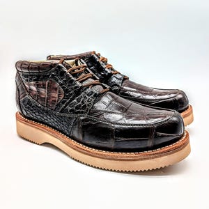 May include: A pair of brown leather boots with a crocodile skin texture. The boots have a rounded toe, a thick, light-colored sole, and brown laces. The boots are approximately 6 inches tall.