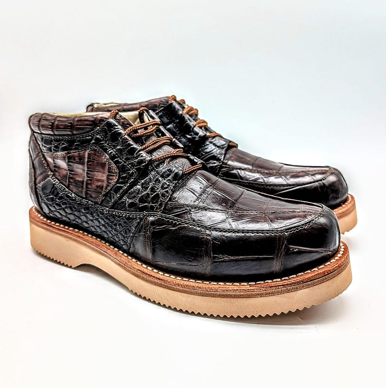 May include: A pair of brown leather boots with a crocodile skin texture. The boots have a rounded toe, a thick, light-colored sole, and brown laces. The boots are approximately 6 inches tall.