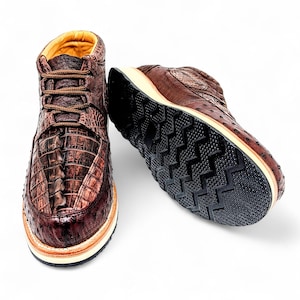 May include: Brown leather high-top trainers with a crocodile texture on the front and a contrasting black sole. The shoes have brown laces and a tan interior. The sole has a unique tread pattern.