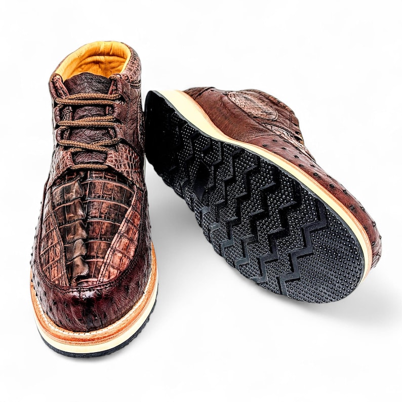 May include: Brown leather high-top trainers with a crocodile texture on the front and a contrasting black sole. The shoes have brown laces and a tan interior. The sole has a unique tread pattern.