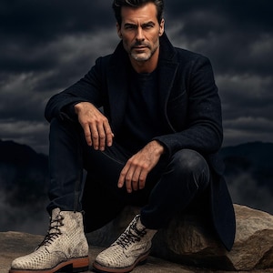 May include: A man in a dark navy coat and jeans, seated on a rock. The image highlights the light-coloured, textured leather boots with brown laces and soles. The boots feature a snakeskin pattern.