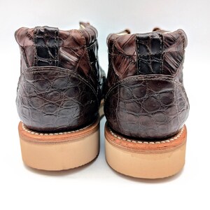 May include: A pair of brown leather boots with a textured, crocodile-like pattern. The boots have a light brown sole and a stitched welt. The uppers are a dark brown color, and the boots are set against a white background.