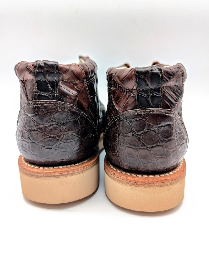 May include: A pair of brown leather boots with a textured, crocodile-like pattern. The boots have a light brown sole and a stitched welt. The uppers are a dark brown color, and the boots are set against a white background.