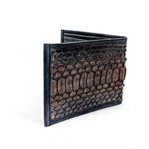 May include: A bi-fold wallet with a brown snakeskin pattern and black trim. The wallet is made of a textured material and has a classic design. The wallet is a stylish accessory for carrying cards and cash.