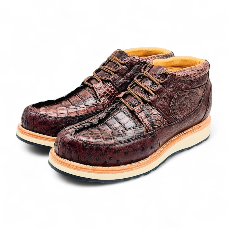 May include: A pair of brown leather boots with a textured crocodile and ostrich pattern. The boots have a lace-up design with brown laces, a beige sole, and a dark brown outsole. The interior lining is yellow.
