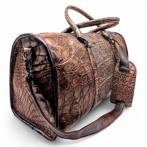 May include: A brown leather duffel bag with a textured, crocodile-like pattern. The bag has a top handle, a shoulder strap, and a luggage tag. The hardware is dark-toned. The bag is designed for travel and has a classic, upscale look.