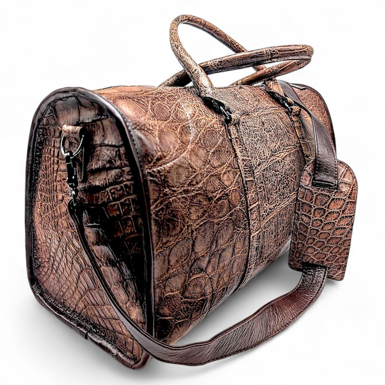 May include: A brown leather duffel bag with a textured, crocodile-like pattern. The bag has a top handle, a shoulder strap, and a luggage tag. The hardware is dark-toned. The bag is designed for travel and has a classic, upscale look.