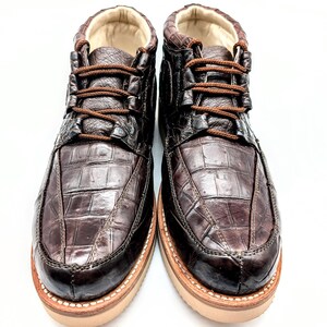 May include: A pair of brown leather boots with a crocodile skin pattern. The boots have brown laces, a tan sole, and a padded interior. The boots are designed for casual wear.