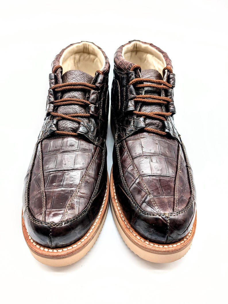 May include: A pair of brown leather boots with a crocodile skin pattern. The boots have brown laces, a tan sole, and a padded interior. The boots are designed for casual wear.