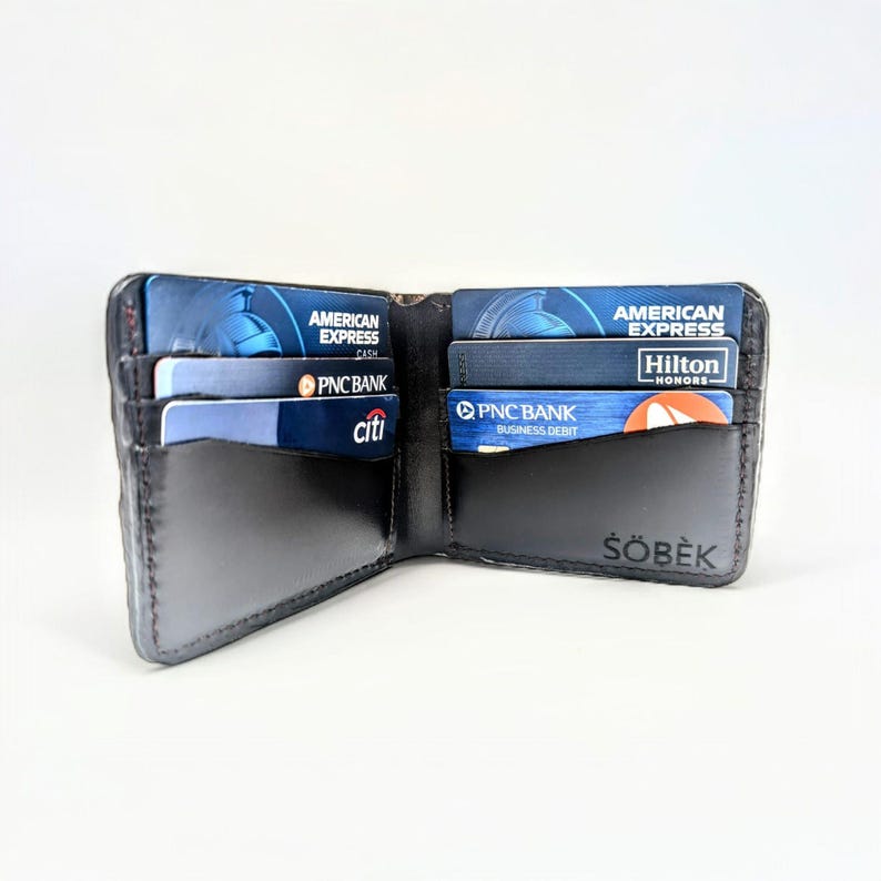 Op de afbeelding: Een zwarte leren portemonnee met meerdere kaartsleuven, open om creditcards te laten zien, waaronder American Express, PNC Bank en Citi. De portemonnee heeft de merknaam "&Scaron;&Ouml;BEK" gegraveerd in de rechterbenedenhoek.