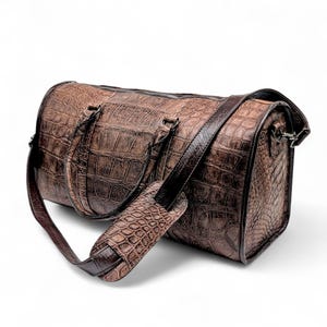 May include: Brown leather duffel bag with a crocodile skin texture. The bag has a cylindrical shape with a zipper closure, two top handles, and a detachable shoulder strap. The bag is made of a dark brown leather.