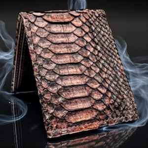 Op de afbeelding: Een bruine bi-fold portemonnee met een slangenleerpatroon. De portemonnee is open, waardoor de binnenkant zichtbaar is. De slangenleertextuur is gedetailleerd en realistisch, met tinten bruin en beige. De achtergrond is donker, met rookpluimen.