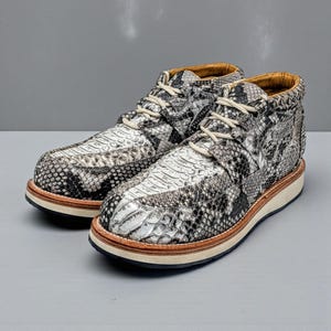 May include: Ankle boots featuring a snakeskin design in grey, black, and white. The boots have beige laces, a tan leather collar, and a light-colored sole with a brown trim. These casual boots are suitable for everyday wear and measure approximately 15 cm in height.