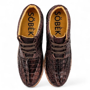 May include: A pair of brown leather shoes with a crocodile skin texture. The shoes have laces and the word "S&Ouml;BEK" is printed in black on the inner sole. The shoes are on a white background.