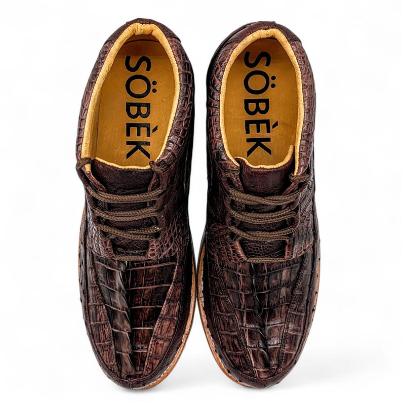 May include: A pair of brown leather shoes with a crocodile skin texture. The shoes have laces and the word "S&Ouml;BEK" is printed in black on the inner sole. The shoes are on a white background.