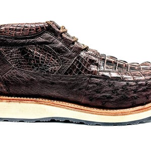 May include: A brown leather boot with a textured crocodile and ostrich pattern. The boot has a thick, off-white sole with a brown trim and black outsole. The laces are brown.