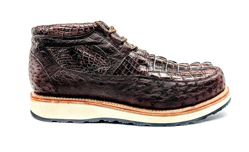 May include: A brown leather boot with a textured crocodile and ostrich pattern. The boot has a thick, off-white sole with a brown trim and black outsole. The laces are brown.