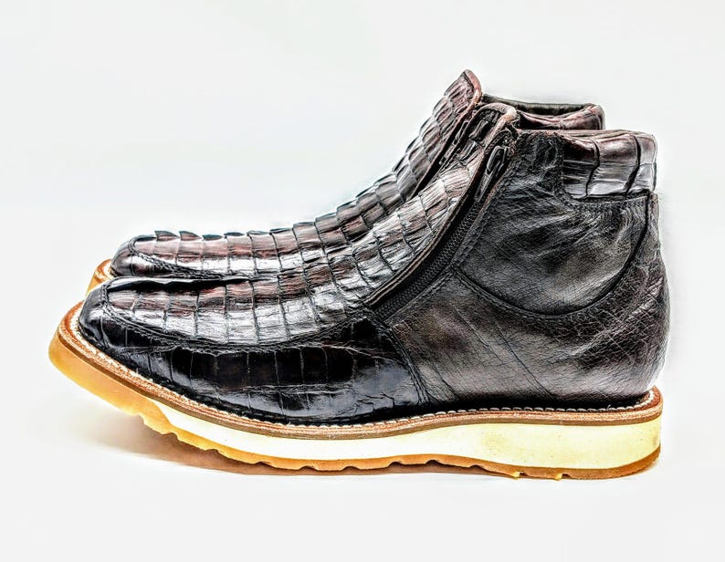 May include: Brown leather boots with a crocodile pattern and a white rubber sole. The boots have a zipper on the side and a pointed toe.