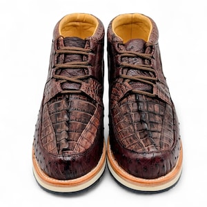 May include: A pair of brown leather ankle boots with a textured, crocodile-like pattern. The boots have brown laces, a tan interior, and a cream and tan sole. The shoes are designed for casual wear.