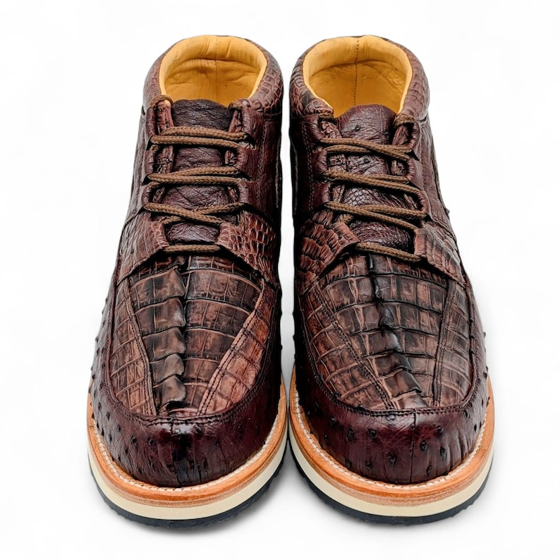 May include: A pair of brown leather ankle boots with a textured, crocodile-like pattern. The boots have brown laces, a tan interior, and a cream and tan sole. The shoes are designed for casual wear.