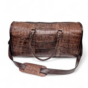 May include: A brown duffel bag with a crocodile skin texture. The bag has a zipper closure, two handles, and a detachable shoulder strap. The bag is made of a durable material and is designed for travel.