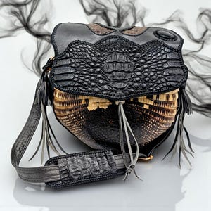May include: A black leather handbag with a crocodile skin flap and a woven basket-like body. The bag has a long, adjustable strap and decorative tassels. The bag is approximately 10 inches wide and 8 inches tall.