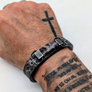 May include: A black and gray leather bracelet with a textured pattern and silver-colored metal clasps. The bracelet is worn on a wrist with a cross tattoo. The bracelet is approximately 0.75 inches wide.
