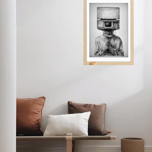May include: Black and white framed art print. The artwork depicts a person with a vintage radio head. The person is wearing a long-sleeved shirt and has their hands clasped in front of them. The art is displayed on a white wall.