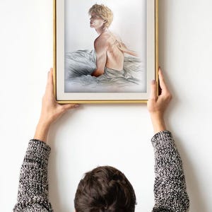 K&ouml;nnte beinhalten: Ein gerahmter Kunstdruck mit einer Bleistiftskizze einer Person mit kurzen blonden Haaren, die auf einem Bett sitzt. Die Person ist zur Seite gedreht, eine Hand ber&uuml;hrt ihren R&uuml;cken. Der Rahmen ist goldfarben.