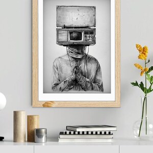 May include: Black and white framed art print. The artwork depicts a person with a vintage television set for a head. The person is wearing a long-sleeved shirt and has their hands clasped in prayer. The art is displayed on a white shelf with books and decorative items.