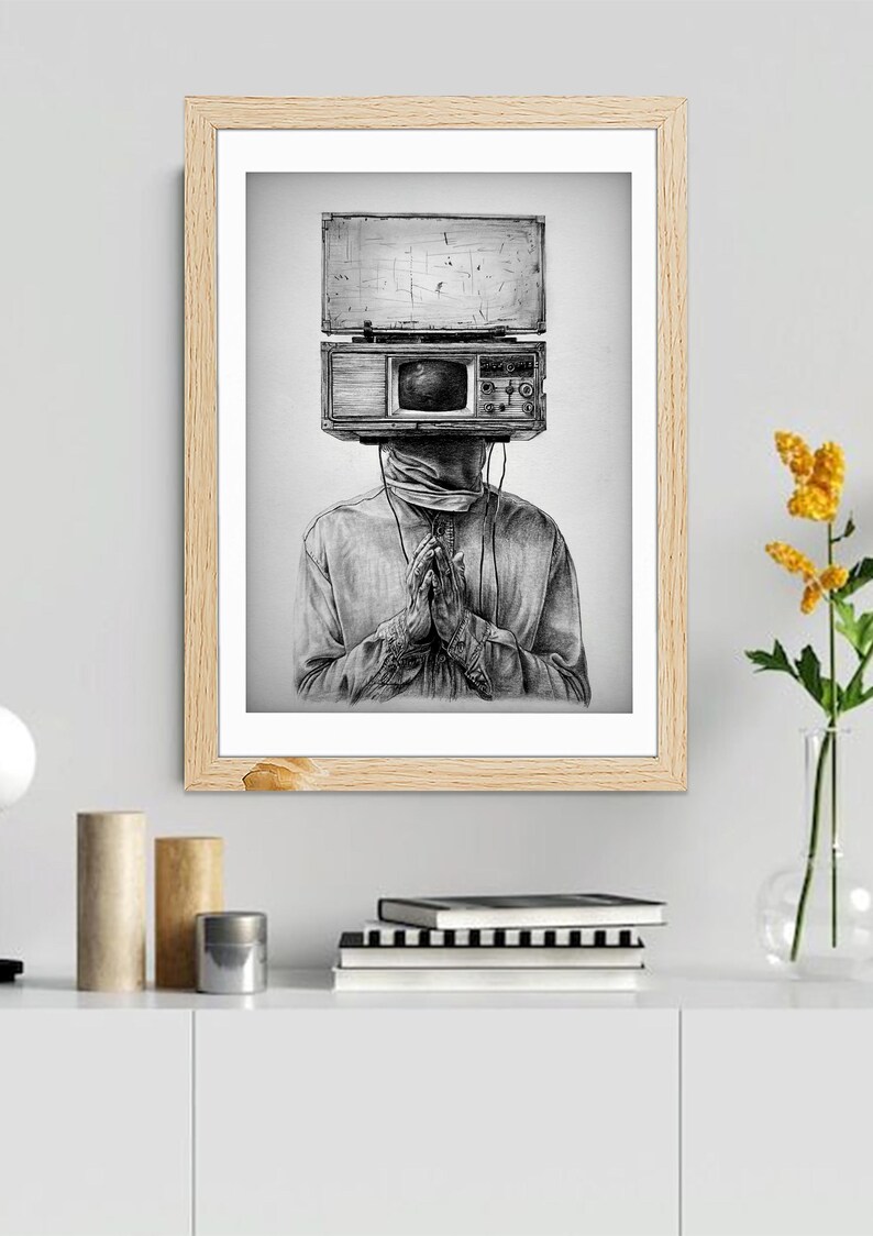 May include: Black and white framed art print. The artwork depicts a person with a vintage television set for a head. The person is wearing a long-sleeved shirt and has their hands clasped in prayer. The art is displayed on a white shelf with books and decorative items.