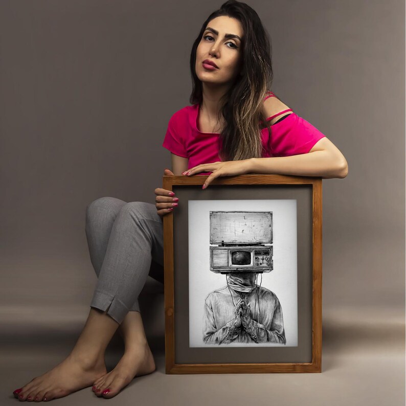 May include: A framed black and white art print featuring a surreal illustration of a person with a vintage radio head. The artwork is held by a person wearing a pink top and gray pants. The frame is brown.