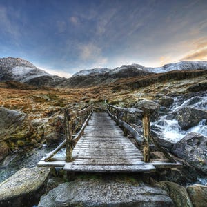 May include: A wooden bridge spans a rocky stream in a mountainous landscape. The bridge is constructed from weathered wooden planks and has wooden handrails. The stream flows over rocks and there is a mountain in the background.