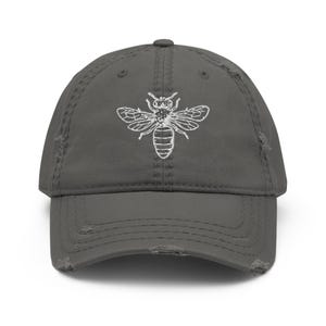 Peut inclure: Casquette de baseball grise délavée avec une broderie blanche d'abeille sur le devant. La casquette a une visière incurvée et un aspect vintage usé, avec des détails effilochés sur les bords.
