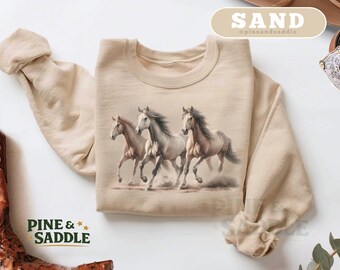 May include: A beige sweatshirt featuring a watercolor illustration of three running horses. The horses are in shades of brown, white, and gray. The text "PINE & SADDLE" is printed in green with yellow stars. The word "SAND" is in a beige rectangle.