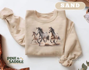 May include: A sand-colored sweatshirt featuring a watercolor illustration of three running horses. The horses are shades of gray and tan, kicking up dust. The text "PINE & SADDLE" is printed in green and gold. The word "SAND" is printed in a tan banner.