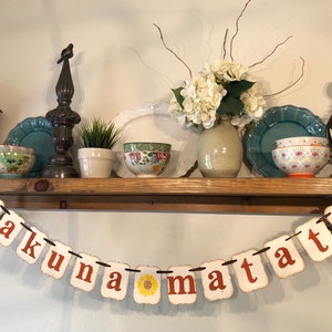 May include: A white banner with the phrase "hakuna matata" in orange letters. The banner is strung with brown twine and is hanging from a wooden shelf. The shelf is decorated with various items, including a vase of white flowers, a small potted plant, and a few bowls.