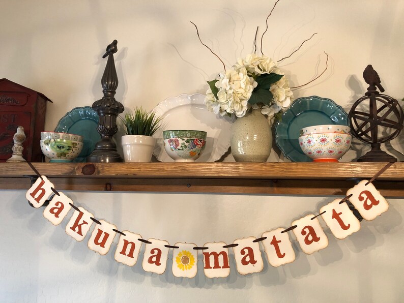 May include: A white banner with the phrase "hakuna matata" in orange letters. The banner is strung with brown twine and is hanging from a wooden shelf. The shelf is decorated with various items, including a vase of white flowers, a small potted plant, and a few bowls.